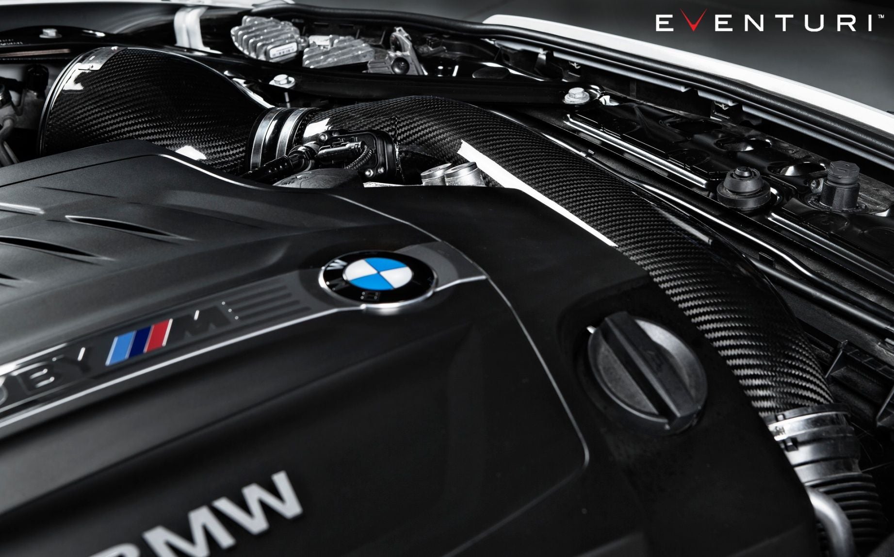 Eine Nahaufnahme eines Motorraums eines BMW M2 F87 mit Kohlefaserkomponenten zeigt deutlich das Eventuri Carbon Ansaugsystem für BMW M2 F87. Das BMW-Logo steht stolz im Vordergrund und fängt den Venturi-Effekt ein. Das Bild strahlt eine elegante, leistungsstarke Ästhetik aus und zeigt Präzisionstechnik und moderne Automobiltechnologie.