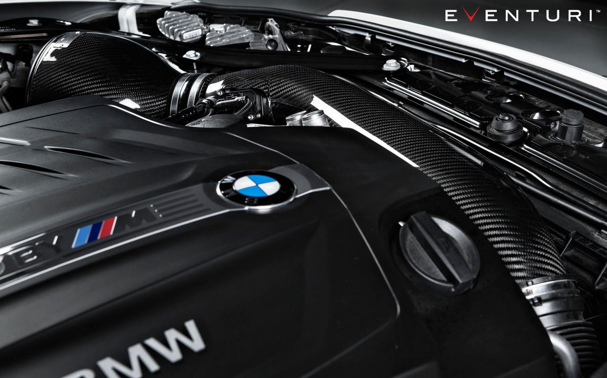 Eine Nahaufnahme eines Motorraums eines BMW M2 F87 mit Kohlefaserkomponenten zeigt deutlich das Eventuri Carbon Ansaugsystem für BMW M2 F87. Das BMW-Logo steht stolz im Vordergrund und fängt den Venturi-Effekt ein. Das Bild strahlt eine elegante, leistungsstarke Ästhetik aus und zeigt Präzisionstechnik und moderne Automobiltechnologie.